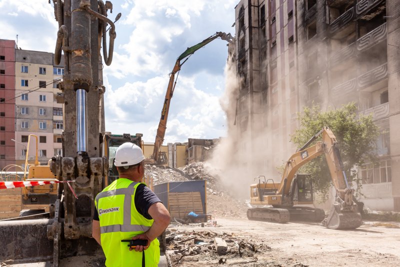 Ein mit neongelber Warnweste und weißem Schutzhelm ausgerüsteter Arbeiter sieht zu, wie im Bildhintergrund ein bombardierter und verbrannter Wohnblock von großen gelben Baggern abgerissen wird. Bei den Abrissarbeiten wird Staub aufgewirbelt.