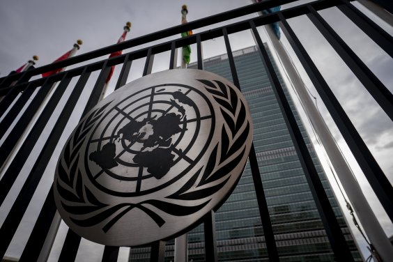 Zu sehen ist das Symbol der Vereinten Nationen am Eingang des UN-Hauptsitzes in New York. Das Symbol zeigt eine Weltkarte umgeben von Olivenzweigen. Im Hintergrund ist das UN-Gebäude, ein Hochhaus, zu erkennen.