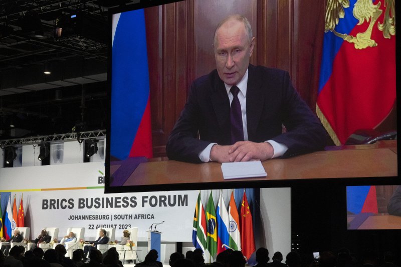 Das Bild zeigt eine Projektion des russischen Präsidenten Wladimir Putin auf einer riesigen Videoleinwand. Unter der Leinwand befindet sich die Bühne des BRICS Business Forum, auf der mehrere Teilnehmerinnen und Teilnehmer sitzen. Sie werden von den Fahnen der BRICS-Mitgliedsstaaten eingerahmt.
