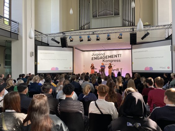Man sieht das Publikum in einem großen Saal von hinten mit Blick auf die Bühne, auf der der Schriftzug "Jugendengagementkongress" zu sehen ist.  