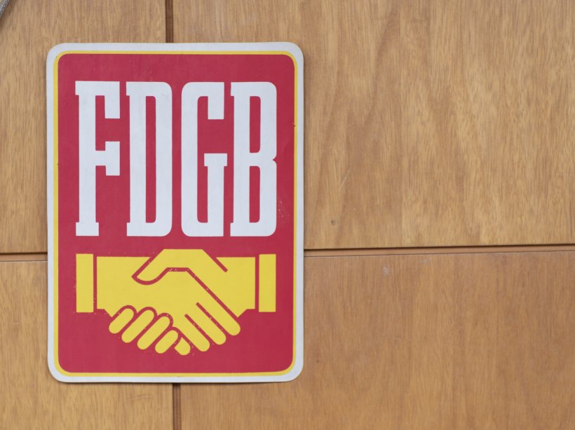 Schild mit Logo des FDGB an einer holzgetäfelten Wand in der Stadthalle Magdeburg. Das Schild zeigt das Kürzel FDGB in weißer Schrift auf rotem Grund, darunter in gelb zwei im Handschlag verbundene Hände.