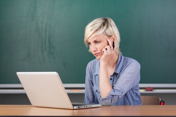 Teaserbild "Webtalkreihe: Schule – Radikalisierung – Prävention" Farbfoto: Eine Frau mit blondem kurzen Haar sitzt vor einer Tafel an einem Schreibtisch. Vor ihr steht ein Laptop, auf den diese Frau schaut.