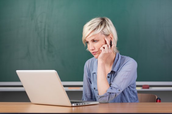 Farbfoto: Eine Frau mit blondem kurzen Haar sitzt vor einer Tafel an einem Schreibtisch. Vor ihr steht ein Laptop, auf den diese Frau schaut. 