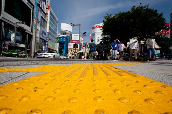 „Eine gelbe Markierung für sehbehinderte Menschen auf dem Boden der Straßen in Tokio. Die mit Noppen und hervorstehenden Linien versehenen Markierungen helfen Blinden durch das lebhafte Stadtviertel Shinjuku zu navigieren