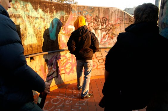 Drei junge Männer gehen eine mit Graffiti beschmierte Treppe herunter. Einer trägt ein Kapuze über den Kopf. Ein anderer hat eine Bierflasche in der Hand. 