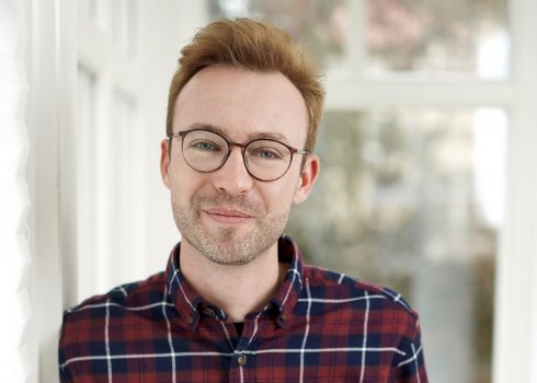 Porträt von Joscha Falck. Er hat kurze blonde Haare, trägt eine Brille und ein kariertes Hemd. Er lächelt in die Kamera. Der Hintergrund ist ein weißes Fenster. Der Hintergrund ist unscharf.