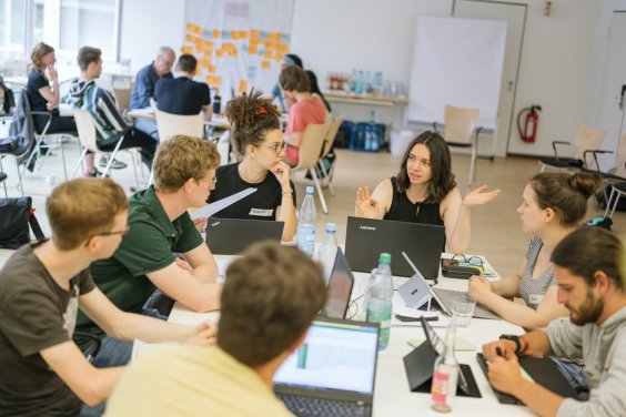 Farbfoto: In einem großen Raum sitzen gemischte Gruppen aus mehreren jungen Frauen und Männer rings um zwei Tische und diskutieren. Im Hintergrund sind viele bunte Klebezettel an einer Wand angebracht. 