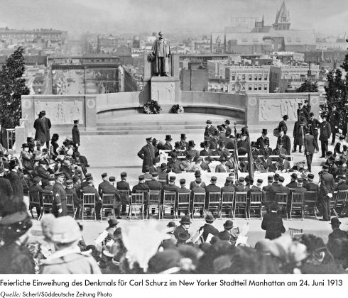 Das Schwarz-weiß-Foto zeigt vor New-Yorker Kulisse eine Menschenmenge von hinten auf Stuhlreihen vor dem Carl-Schurz-Denkmal, das mit Blumenkränzen geschmückt ist. Die Menge besteht überwiegend aus Männern, teils in Uniform, teils im schwarzen Anzug mit Hut. Die wenigen Frauen tragen Kleider und Hüte.