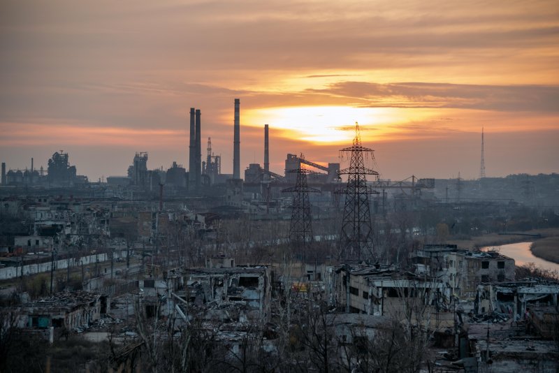 Das Bild zeigt die Ruinen zerstörter Gebäude vor dem Asovstal-Stahlwerk in Mariupol. Das Stahlwerk selbst ist im Sonnenuntergang in Sillouettenform zu erkennen.